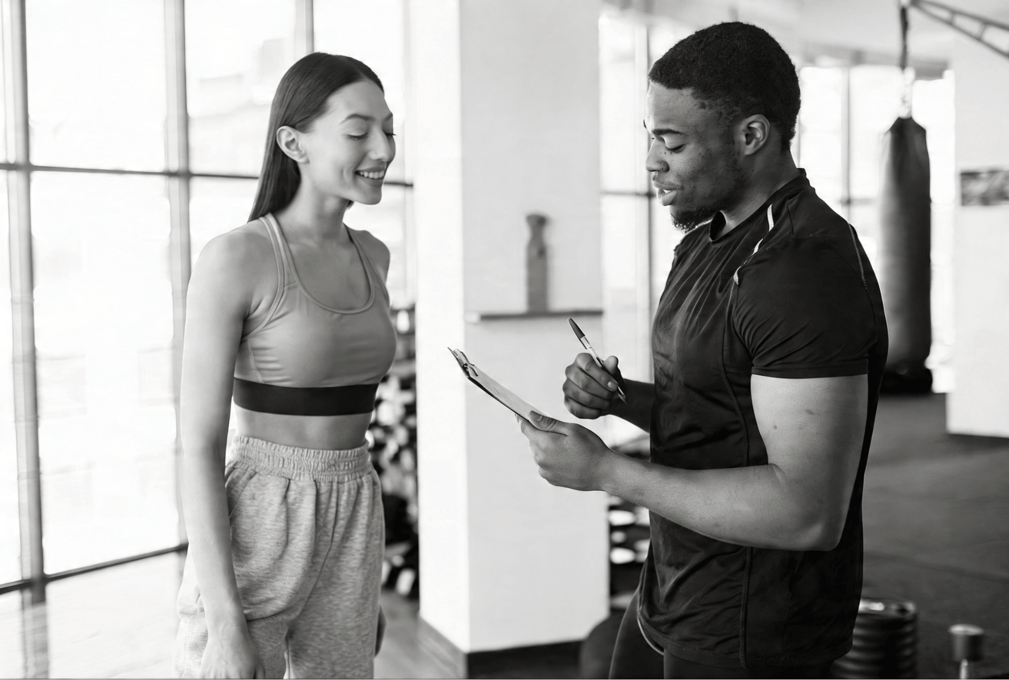image1 Black-and-white photo of a trainer or coach holding a clipboard and talking with a smiling athlete in a gym.