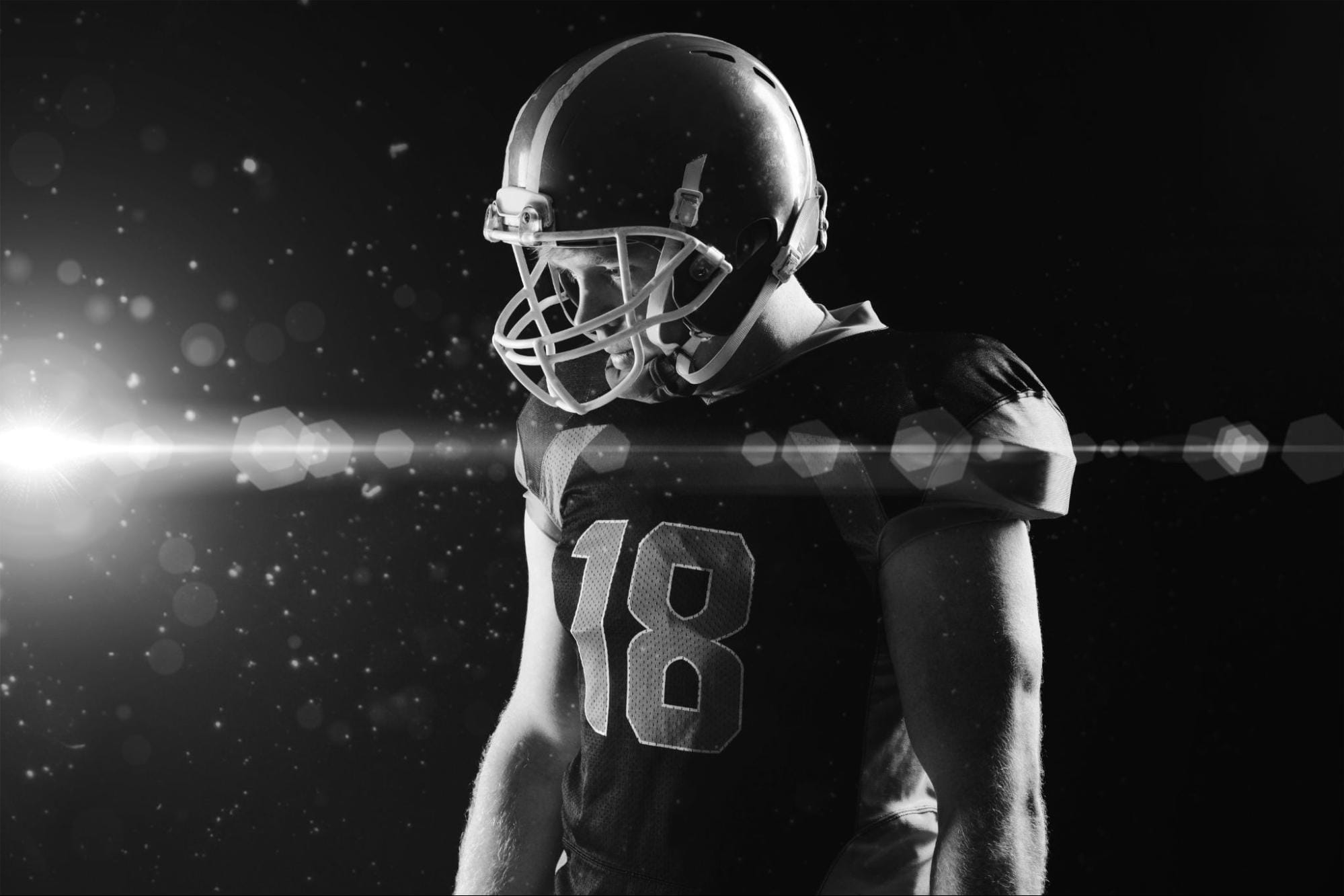 image1 Black-and-white photo of a football player in uniform and helmet standing under dramatic lighting against a dark background.